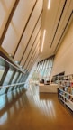 An interior space featuring an architecturally modern design with angled walls and large windows. The area is well-lit with a smooth, reflective brown floor. Shelving units display various items and a desk area is situated at the end of the space.