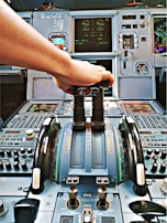 A cockpit control panel with various switches, dials, and screens. A hand is reaching forward to adjust a lever, likely the throttle. The control panel is intricate, featuring numerous gauges, digital displays, and buttons.