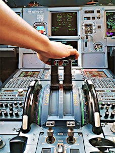 A cockpit control panel with various switches, dials, and screens. A hand is reaching forward to adjust a lever, likely the throttle. The control panel is intricate, featuring numerous gauges, digital displays, and buttons.