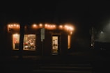 A warmly lit shop at night with strings of lights hanging from the roof. Inside, a person sits in a chair facing a cluttered shelf, which is visible through a large window. The shop is adorned with a sign, and a street sign is positioned nearby. There is a faint glow of light shining through the fog outside.
