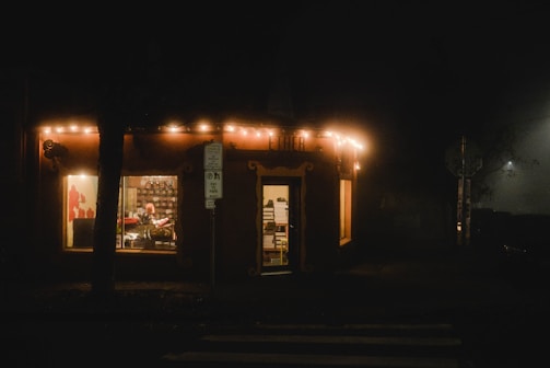 A warmly lit shop at night with strings of lights hanging from the roof. Inside, a person sits in a chair facing a cluttered shelf, which is visible through a large window. The shop is adorned with a sign, and a street sign is positioned nearby. There is a faint glow of light shining through the fog outside.