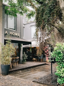 An inviting outdoor seating area of a cafe or restaurant is adorned with lush greenery and flowering plants, creating a cozy atmosphere. Wooden furniture and potted plants are scattered around the space, with soft lighting visible inside through large windows.