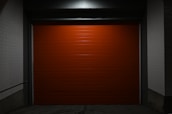 A closed, bright red garage door centered within a dimly lit concrete structure. The surrounding walls are painted white, and the scene is enveloped in shadows, creating a stark contrast against the vibrant door.
