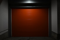 A closed, bright red garage door centered within a dimly lit concrete structure. The surrounding walls are painted white, and the scene is enveloped in shadows, creating a stark contrast against the vibrant door.