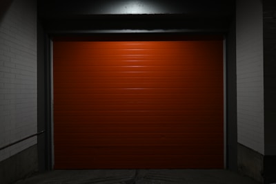 A closed, bright red garage door centered within a dimly lit concrete structure. The surrounding walls are painted white, and the scene is enveloped in shadows, creating a stark contrast against the vibrant door.