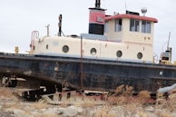An old and weathered boat, likely retired from active service, is resting on land surrounded by dry vegetation. The boat is cream and red with some rust patches and has multiple circular windows along its side. It appears to be a work vessel, possibly used by the US Army Corps of Engineers, indicated by a visible sign on the side.