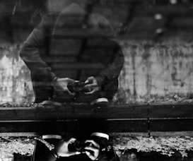 A reflective image captures a person holding a camera, with their reflection in water creating a mirror effect. The black and white tones add a moody atmosphere, and the reflection blurs the distinction between the person and their surroundings.