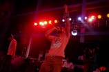A performer passionately sings into a microphone under vibrant stage lighting with red and pink hues. The spotlight is on the singer, who is energetically raising one arm. Other musicians and people appear in the background, contributing to a lively concert atmosphere.
