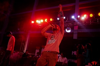 A performer passionately sings into a microphone under vibrant stage lighting with red and pink hues. The spotlight is on the singer, who is energetically raising one arm. Other musicians and people appear in the background, contributing to a lively concert atmosphere.