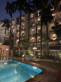 A grand hotel building stands illuminated at night, surrounded by tall palm trees. The structure features multiple balconies with warm-lit windows. A swimming pool with clear blue water reflects the lights and the archways along its perimeter. Outdoor seating with tables and chairs is arranged neatly on the patio, adding to the elegant ambiance.