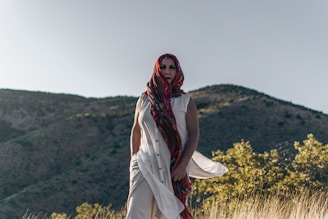 A person wearing a colorful, patterned headscarf and a flowing white garment stands in a natural setting with mountains in the background and dry grasses in the foreground.