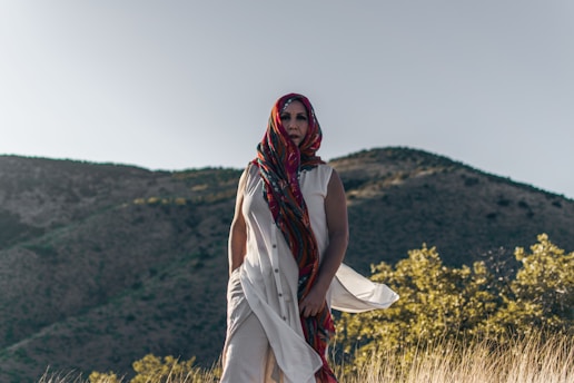 A person wearing a colorful, patterned headscarf and a flowing white garment stands in a natural setting with mountains in the background and dry grasses in the foreground.