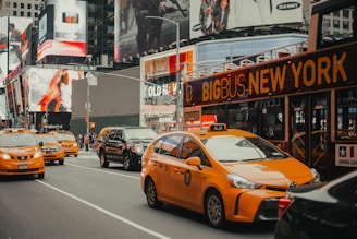 A busy urban street scene featuring yellow taxis driving alongside a double-decker bus with 'Big Bus New York' signage. Surrounding buildings are adorned with large advertisements and electronic billboards, typical of a bustling city atmosphere.