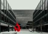A modern glass office building with a symmetrical design featuring a prominent red abstract sculpture in the foreground. The structure has multiple floors with uniform window panels, and the courtyard includes small trees and paved walkways.