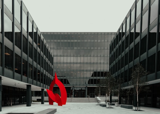 A modern glass office building with a symmetrical design featuring a prominent red abstract sculpture in the foreground. The structure has multiple floors with uniform window panels, and the courtyard includes small trees and paved walkways.