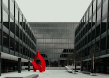A modern glass office building with a symmetrical design featuring a prominent red abstract sculpture in the foreground. The structure has multiple floors with uniform window panels, and the courtyard includes small trees and paved walkways.