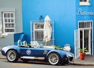A vintage blue sports car is parked on the street in front of a blue building with the name 'Batavia Cafe' written on it. The building has white barred windows and a white door, with several potted plants surrounding the entrance. Closed white umbrellas are positioned near the windows, and the scene has a quaint, classic ambiance.
