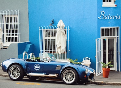 A vintage blue sports car is parked on the street in front of a blue building with the name 'Batavia Cafe' written on it. The building has white barred windows and a white door, with several potted plants surrounding the entrance. Closed white umbrellas are positioned near the windows, and the scene has a quaint, classic ambiance.