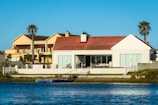 A waterfront scene featuring two large, modern houses with spacious terraces facing a calm body of water. The houses have palm trees nearby, adding a tropical feel to the environment. The sky is clear and blue, and the water reflects the buildings and the sky.