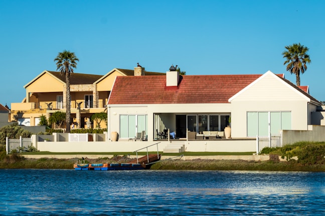 A waterfront scene featuring two large, modern houses with spacious terraces facing a calm body of water. The houses have palm trees nearby, adding a tropical feel to the environment. The sky is clear and blue, and the water reflects the buildings and the sky.