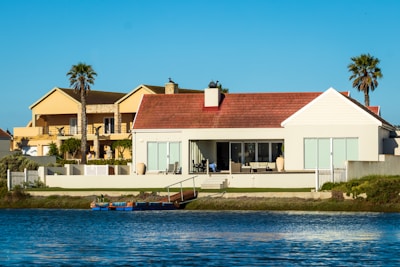 A waterfront scene featuring two large, modern houses with spacious terraces facing a calm body of water. The houses have palm trees nearby, adding a tropical feel to the environment. The sky is clear and blue, and the water reflects the buildings and the sky.