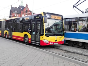 A public bus with a destination sign reading 'ZWYCIESKA' is stopped on a street. The bus is predominantly yellow with red and black accents and has the number 8420 displayed on it. Beside the bus, there is a tram in blue and white colors. The background includes a brick building with green spires and several windows. The street is paved with stone tiles.