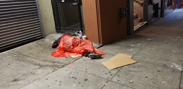 A person is lying on the sidewalk covered with an orange tarp or blanket, situated near a closed storefront with a metal shutter. Nearby are some scattered items, including a cardboard piece and a pair of shoes. The area appears to be dimly lit, suggesting nighttime.