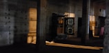 A dimly lit industrial interior with concrete pillars and walls, featuring an open vault door revealing shelves inside. Warm light casts shadows and highlights across the rusticated surfaces, creating a moody and mysterious atmosphere.