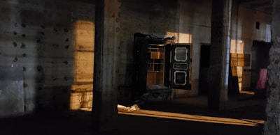 A dimly lit industrial interior with concrete pillars and walls, featuring an open vault door revealing shelves inside. Warm light casts shadows and highlights across the rusticated surfaces, creating a moody and mysterious atmosphere.