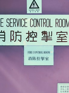The image features a pinkish-purple wall with three prominent signs. The first sign at the top contains a warning about electrical hazards, featuring a lightning bolt symbol and text in both English and Chinese. Below this, a larger sign reads 'FIRE SERVICE CONTROL ROOM' in both English and Chinese. At the bottom, a smaller sign includes additional text, also in Chinese.