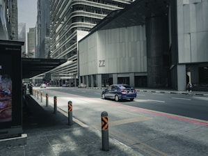 A city street with modern high-rise buildings, featuring a blue car driving along a quiet road. The area appears to be urban with tall structures and a shadowed bus stop area on the sidewalk. The sunlight casts direct light onto the street, highlighting the road and the car.