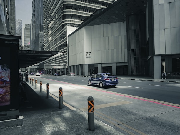 A city street with modern high-rise buildings, featuring a blue car driving along a quiet road. The area appears to be urban with tall structures and a shadowed bus stop area on the sidewalk. The sunlight casts direct light onto the street, highlighting the road and the car.