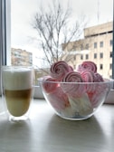A cozy kitchen scene featuring a bowl filled with pink and white meringue cookies styled like rose flowers, placed next to a glass mug of layered coffee with frothed milk. Both items rest on a white wooden tabletop against the backdrop of a window showing a blurry view of bare trees and buildings.