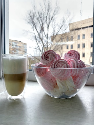 A cozy kitchen scene featuring a bowl filled with pink and white meringue cookies styled like rose flowers, placed next to a glass mug of layered coffee with frothed milk. Both items rest on a white wooden tabletop against the backdrop of a window showing a blurry view of bare trees and buildings.