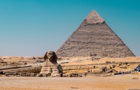 A vast desert landscape with the iconic Great Sphinx and a pyramid in the background. The Sphinx, a monumental limestone statue with a lion's body and a human head, sits prominently in the foreground. Behind it, the pyramid rises, constructed from large stone blocks. Scattered visitors and tourists explore the area, giving a sense of scale to the massive structures. The sky is a clear blue, contrasting with the sandy tones of the desert and monuments.