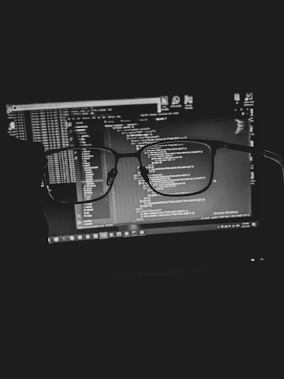 A pair of eyeglasses is held in front of a laptop screen displaying lines of code. The image is in black and white, giving a focused and professional atmosphere. The code is slightly blurred through the lens of the glasses, suggesting a theme related to programming or coding focus.