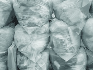 A collection of large, tightly sealed, transparent plastic bags filled with various items, stacked neatly on top of each other. The contents within the bags include #B8C0CC objects and waste material, creating a textured surface. The overall scene conveys a sense of organized clutter.