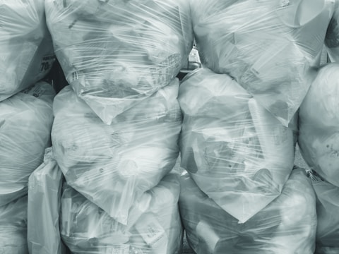 A collection of large, tightly sealed, transparent plastic bags filled with various items, stacked neatly on top of each other. The contents within the bags include #B8C0CC objects and waste material, creating a textured surface. The overall scene conveys a sense of organized clutter.