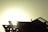 A silhouetted figure of a person is seen working on the roof of a structure against the backdrop of a bright sun. The structural components are clearly visible, with beams and supports highlighted by the strong light source.