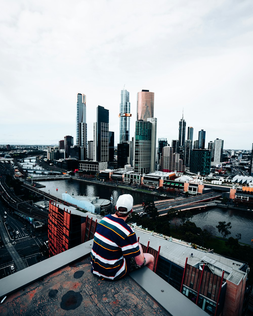 man in white red and blue striped shirt sitting on the edge of a building looking