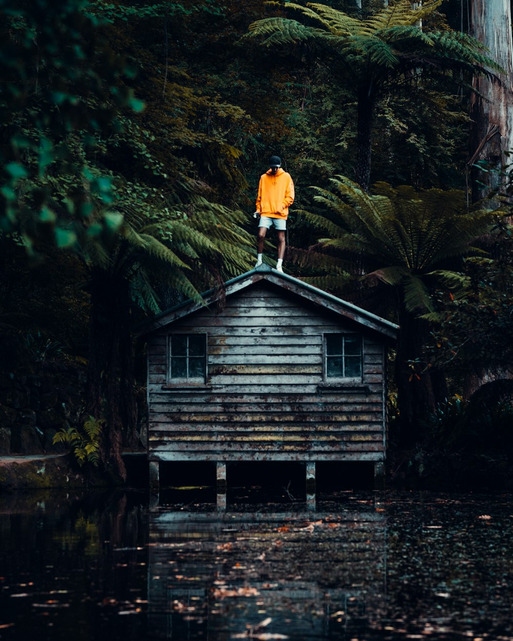 man in orange jacket standing on brown wooden house in the middle of the forest