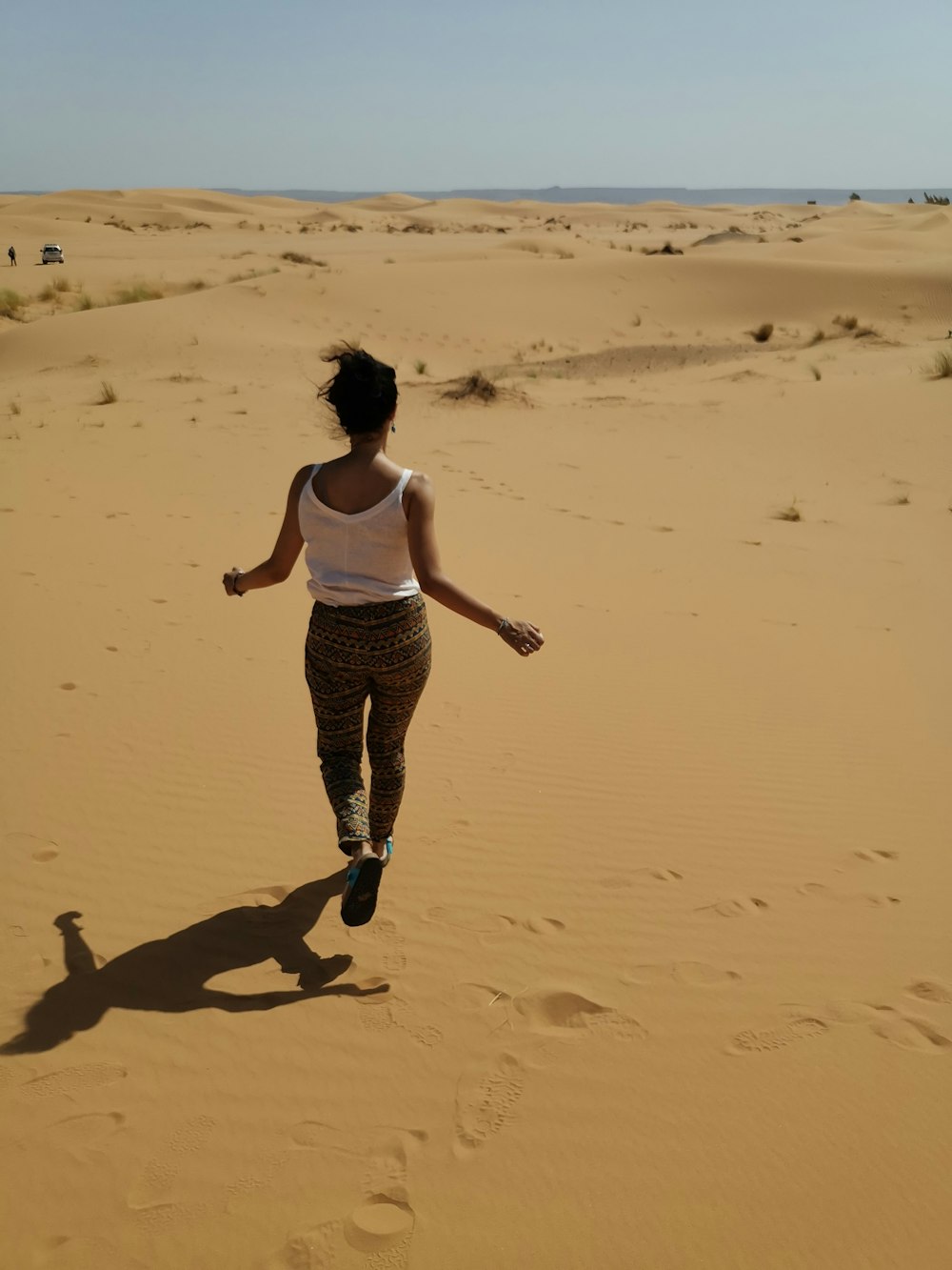 woman in white tank top and black and white pants walking on brown sand during daytime