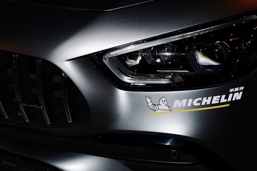 Close-up view of a car's front section, highlighting the headlight and part of the grille. A Michelin sticker is prominently displayed on the car's body, featuring the recognizable Michelin Man logo.
