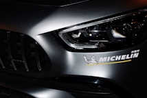 Close-up view of a car's front section, highlighting the headlight and part of the grille. A Michelin sticker is prominently displayed on the car's body, featuring the recognizable Michelin Man logo.