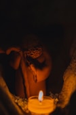 A small, intricately designed clay figure is placed in a dimly lit environment. In front of it, a candle provides a warm glow, accentuating the earthy tones of the statue. The figure appears to be holding an object or gesture towards the light, creating a serene and contemplative atmosphere.