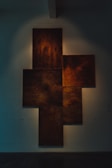 A series of overlapping rectangular panels with a textured, rust-like appearance are mounted on a wall. They are illuminated with soft, dramatic lighting that highlights the warm, earthy tones. The arrangement is symmetrical and artistic, creating an abstract and geometric form.