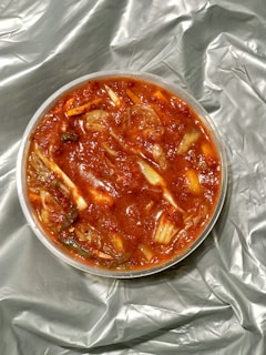 A round container filled with a chunky, vibrant red dish, resembling kimchi. Ingredients include visible cabbage pieces and possibly spices, immersed in a rich, red sauce. The container rests on a crumpled, silver-gray fabric or material.