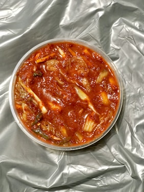 A round container filled with a chunky, vibrant red dish, resembling kimchi. Ingredients include visible cabbage pieces and possibly spices, immersed in a rich, red sauce. The container rests on a crumpled, silver-gray fabric or material.