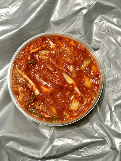 A round container filled with a chunky, vibrant red dish, resembling kimchi. Ingredients include visible cabbage pieces and possibly spices, immersed in a rich, red sauce. The container rests on a crumpled, silver-gray fabric or material.