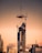 A high-rise building under construction with a crane lifting a large metal platform at the top. The structure is surrounded by other tall buildings, and the sky is a warm gradient with a golden orange hue, indicating either sunrise or sunset.